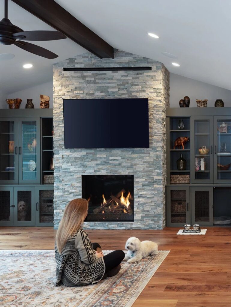 Stone fireplace surrounded by gray built-in cabinetry with a homeowner and her pup on a rug
