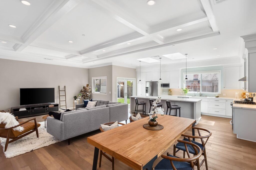 Open-concept living area featuring a wooden dining table in the foreground, a gray sectional sofa facing a television, and a kitchen with a white island and bar seating.