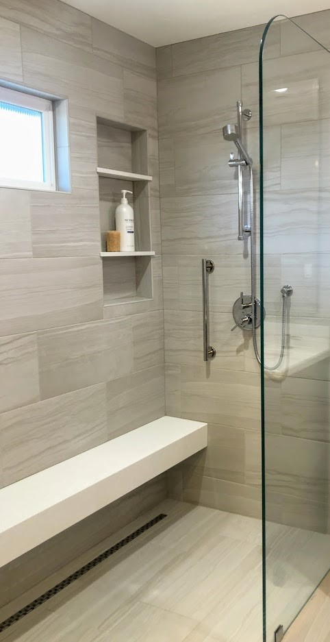 Walk-in shower with light gray tiled walls, a built-in white bench, a glass partition, and a recessed wall niche holding a bottle of soap and a sponge.
