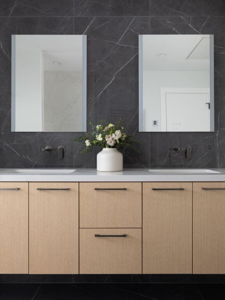 Double vanity with light wood cabinets, two rectangular mirrors, wall-mounted faucets, and a flower arrangement centered on a white countertop against a dark marble wall.