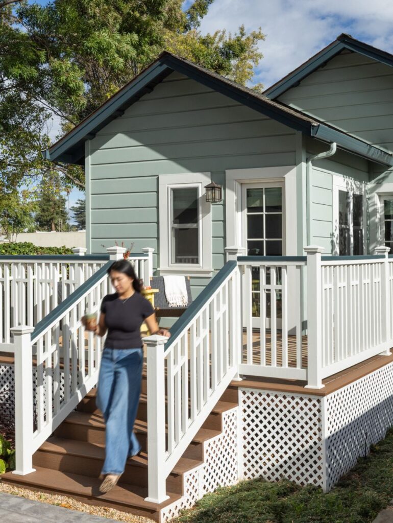 A woman walking down the steps of a light green house with white railing and a wooden deck.