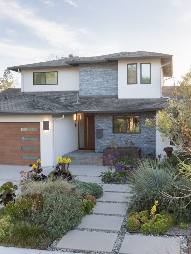 A two-story house with white stucco, gray stone siding, and a wooden garage door, featuring a stone path leading to the front entrance through a front garden.