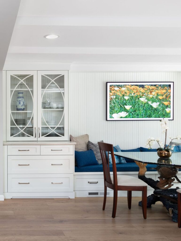 A white dining hutch stands next to a built-in bench with blue cushions, a wooden chair, and a dining table in front of a wall-mounted television displaying a field of yellow flowers.