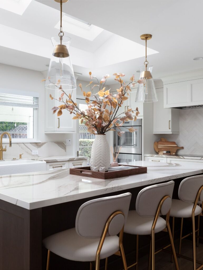 A vase of autumn-colored branches sits on a white marble kitchen island featuring three upholstered stools with gold frames.